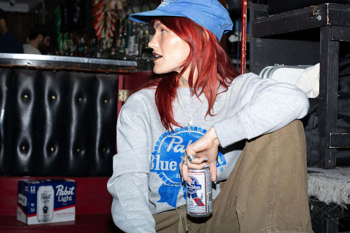 Person sitting in a bar holding a beer, wearing a blue cap and gray sweatshirt with 'Pabst Blue Ribbon' branding.